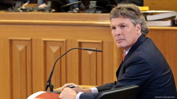 Ein Mann im Anzug sitzt an einem Tisch mit Mikrofon und blickt zur Seite.