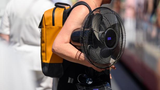 Symboldbild: Eine Person trägt einen gelben Rucksack und einen schwarzen Tischventilator der Marke SMC unter dem Arm.