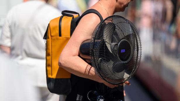 Symboldbild: Eine Person trägt einen gelben Rucksack und einen schwarzen Tischventilator der Marke SMC unter dem Arm.