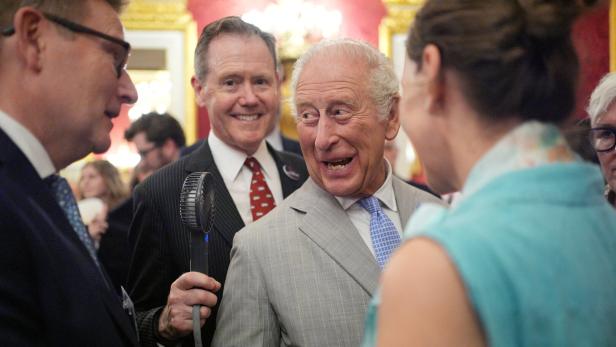König Charles III. unterhält sich in einem Raum mit Gästen, während ein Mann im Hintergrund einen kleinen Handventilator hält.
