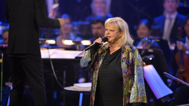 Symbolbild: Sängerin Marianne Mendt in einer glitzernden Jacke singt mit einem Mikrofon vor einem Orchester.
