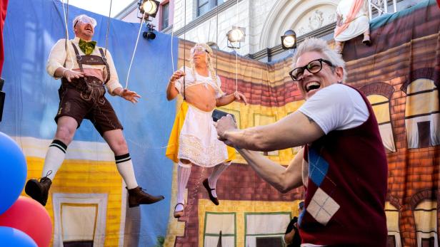 Johnny Knoxville filmt zwei an Seilen hängende Männer in bayerischer Tracht und Dirndl vor einer bemalten Kulisse.