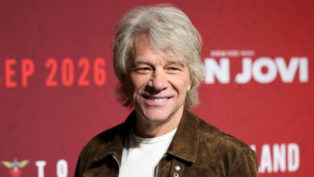 Jon Bon Jovi lächelt vor einem roten Hintergrund mit dem Schriftzug „Bon Jovi“.
