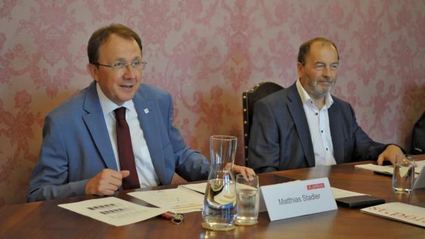 Zwei Männer im Anzug sitzen an einem Konferenztisch mit Wasserkrügen und Namensschild.