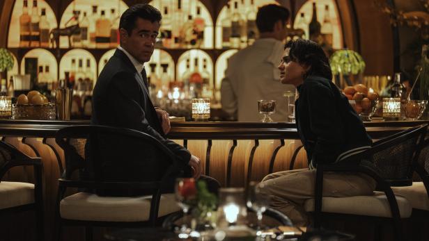 Colin Farrell im Anzug und eine Frau sitzen sich an einer beleuchteten Bar gegenüber und unterhalten sich.