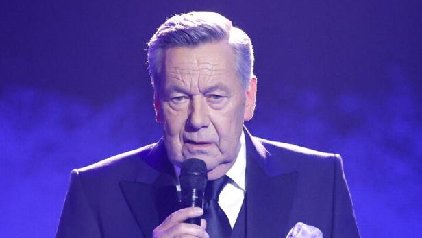 Roland Kaiser steht in einem dunklen Anzug mit Krawatte und Einstecktuch vor einem blauen Hintergrund und spricht in ein Mikrofon.