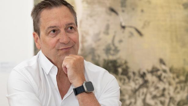 Dieter Nuhr in einem weißen Hemd stützt sein Kinn auf die Hand und blickt zur Seite vor einem abstrakten, hellen Hintergrund.