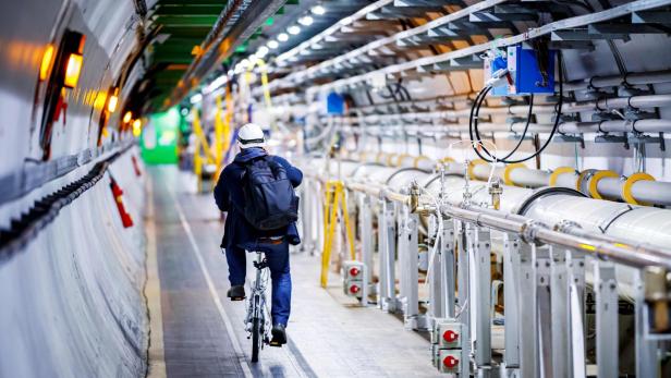 Eine Person mit Helm und Rucksack fährt auf einem Fahrrad durch einen langen, technisch ausgestatteten Tunnel des CERN.