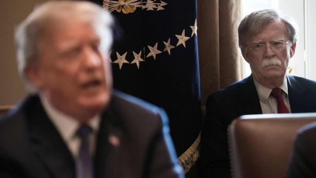 Donald Trump spricht unscharf im Vordergrund, während John Bolton im Hintergrund vor einer US-Präsidentenflagge sitzt.