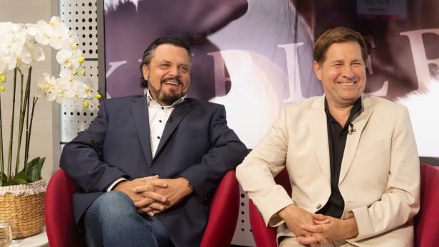 Werner Auer und Erich Furrer im Gespräch sitzen lachend in roten Sesseln, der eine in dunklem, der andere in hellem Anzug.
