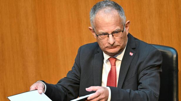Markus Marterbauer liest in einer Mappe an einem Tisch vor braunem Hintergrund.