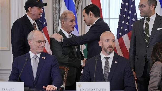 Marco Rubio begrüßt einen Mann vor Flaggen der USA und Israels, während weitere Personen an Tischen mit Namensschildern sitzen.