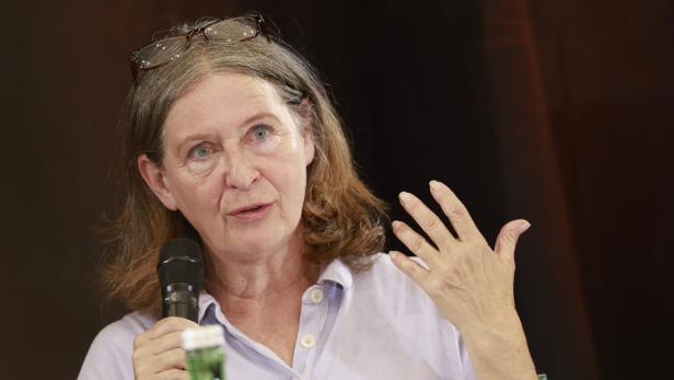 Eine Frau mit Brille auf dem Kopf spricht in ein Mikrofon und gestikuliert mit der Hand vor einem dunklen Hintergrund.