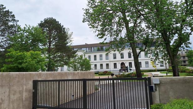 Ein modernes weißes Gebäude hinter einem schwarzen Tor, umgeben von hohen Bäumen und Grün.
