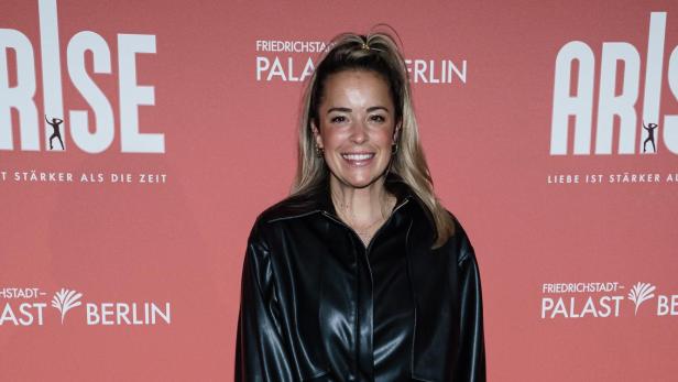 Lächelnde Frau in schwarzem Leder-Outfit mit grüner Handtasche vor pinker ARISE-Wand in Berlin.