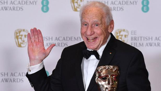 Mel Brooks lächelt fröhlich und winkt vor einer Kulisse der EE British Academy Film Awards mit einer BAFTA-Trophäe in der Hand.