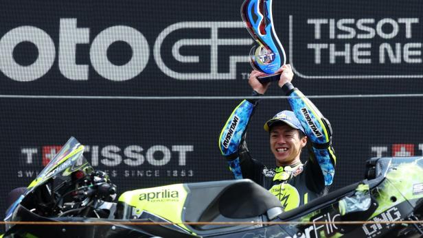 Ai Ogura feiert mit einer blauen Trophäe in der Hand. Vor ihm steht sein neongelb-schwarzes Motorrad. 