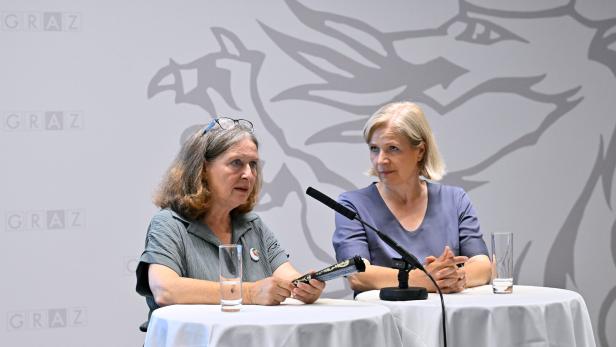 Zwei Frauen sitzen vor einem Hintergrund mit dem Grazer Stadtwappen an Stehtischen bei einer Pressekonferenz.