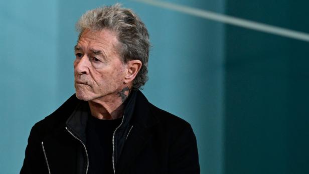 Peter Maffay mit grauem Haar und einem Tattoo am Hals vor einem türkisfarbenen Hintergrund.