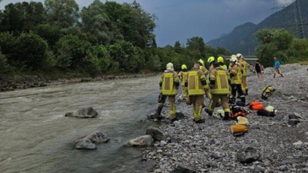 Feuerwehrleute in Schutzkleidung stehen am Ufer eines Flusses unter dunklen Wolken und bereiten Ausrüstung vor.