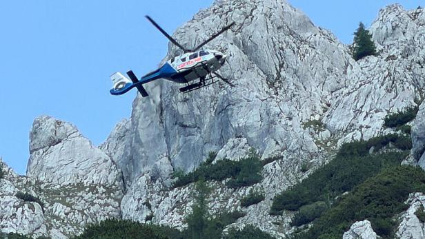 Hubschrauber fliegt über einer felsigen Berglandschaft mit wenigen Bäumen.