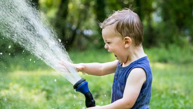 Ein kleiner Junge spritzt im Garten mit einem Gartenschlauch Wasser in die Luft.