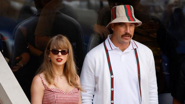 Taylor Swift und Travis Kelce sitzen nebeneinander auf einer Tribüne, wobei sie ein rot-weiß kariertes Kleid und er einen Gucci-Hut trägt.