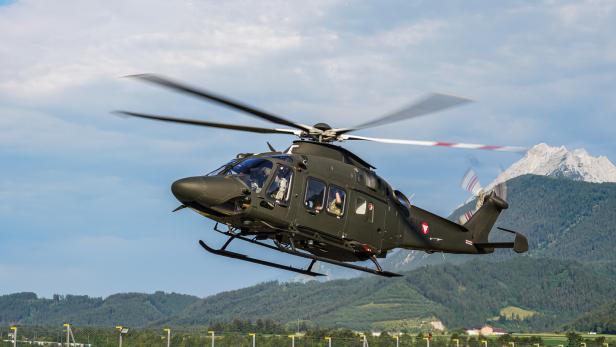 Ein olivgrüner Hubschrauber des Österreichischen Bundesheeres fliegt vor einer Bergkulisse.