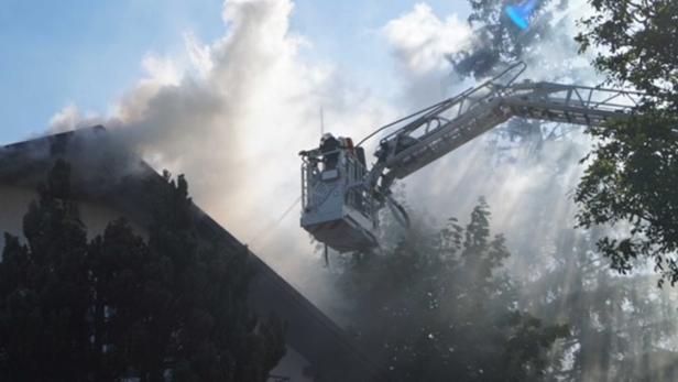 Feuerwehr löscht Brand an einem Dach mit Drehleiter, Rauch steigt auf.