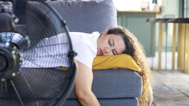 Eine Frau schläft erschöpft auf einer Couch, ihr Kopf liegt auf einem gelben Kissen, davor ein laufender Ventilator.