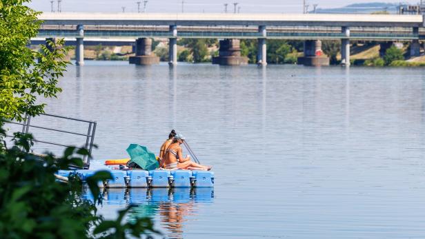 Zwei Frauen sitzen in Badekleidung auf einem blauen Schwimmsteg im Wasser vor einer großen Brücke.