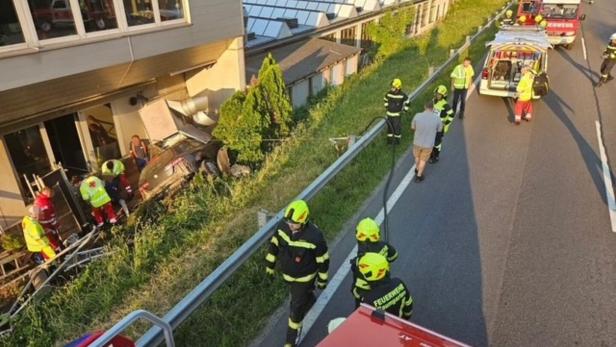 Feuerwehrleute und Rettungskräfte bergen ein Auto an einem Hang neben einem Gebäude; mehrere Einsatzfahrzeuge stehen auf der Straße.