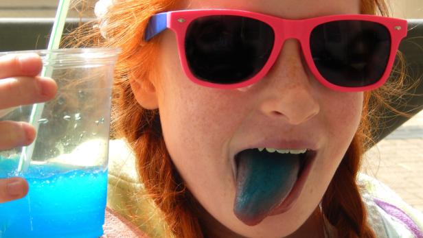 Ein Mädchen hält einen blauen Slushie in der Hand und streckt eine blaue Zunge heraus.
