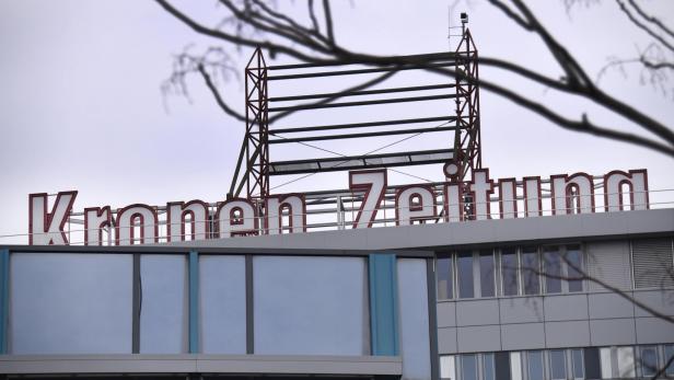 Dach eines Gebäudes mit dem Schriftzug „Kronen Zeitung“ vor grauem Himmel und kahlen Ästen.