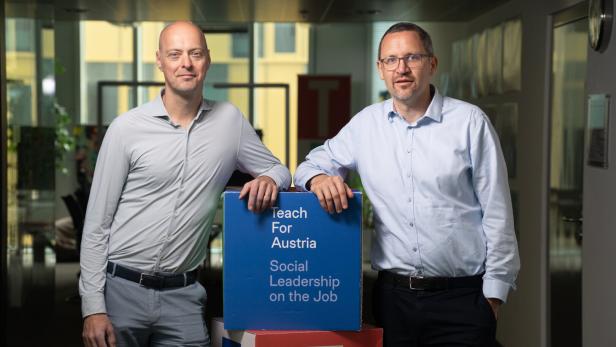 Zwei Männer stehen in einem Büroflur neben einem blauen Würfel mit der Aufschrift „Teach For Austria“.
