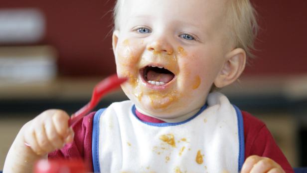 Ein lachendes Baby mit Brei verschmiertem Gesicht hält einen Löffel.