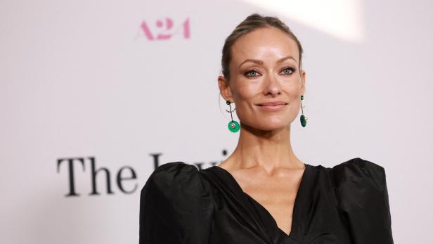 Olivia Wilde trägt ein schwarzes Oberteil und grüne Ohrringe vor einem A24-Hintergrund.