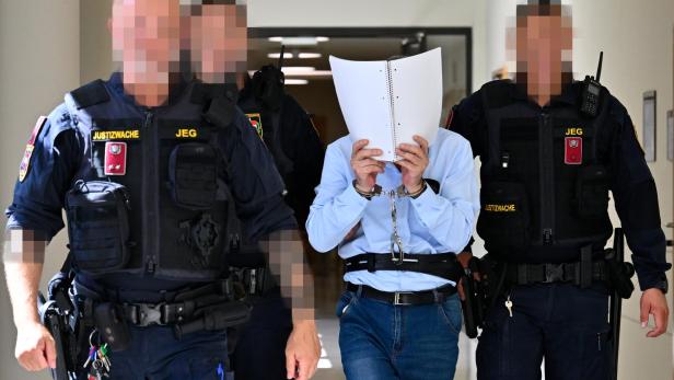 Handschellen Mann mit Aktenblättern vor dem Gesicht, begleitet von drei Justizwachen in einem Flur.