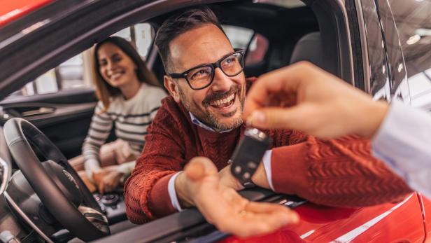 Mann im Auto übergibt lachend einen Autoschlüssel an eine Person draußen, Frau sitzt auf dem Rücksitz.