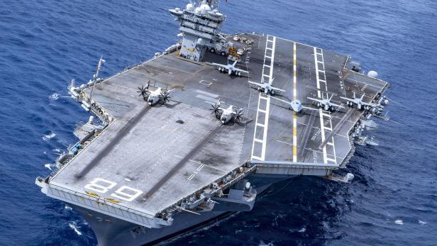 Ein Flugzeugträger der US Navy mit der Kennung 89 fährt auf dem offenen Meer, während mehrere Militärflugzeuge auf seinem Deck geparkt sind.