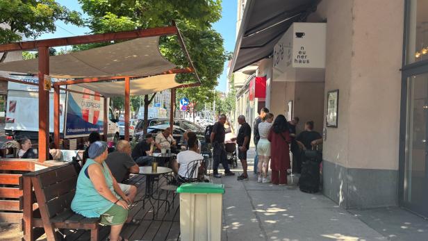 Menschen sitzen vor dem Café Neunerhaus auf einer Terrasse unter einem Sonnensegel, während andere vor dem Eingang warten.