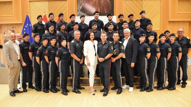 Polizeigruppe in Uniform posiert mit zwei Zivilpersonen in einem Saal vor EU- und Österreich-Flaggen.