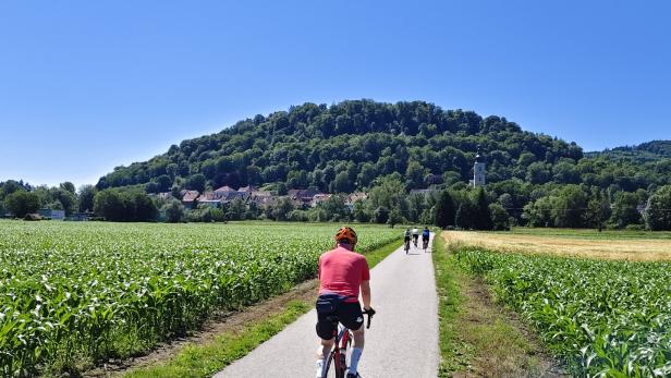 Eine Gruppe von Radfahrern fährt auf einem asphaltierten Weg zwischen Feldern auf ein Dorf mit bewaldetem Hügel zu.