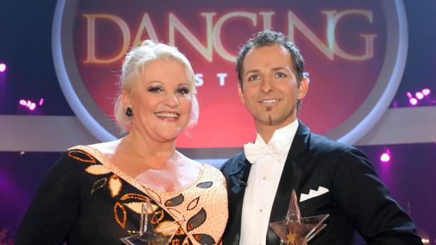 Marika Lichter und ihr Tanzpartner präsentieren stolz ihre Trophäen vor dem Logo der ORF-Show Dancing Stars.
