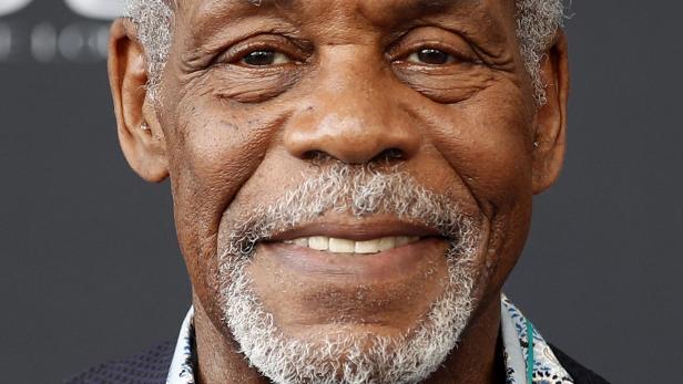 Nahaufnahme des lächelnden Schauspielers Danny Glover mit grauem Haar und Bart vor einem dunklen Hintergrund.