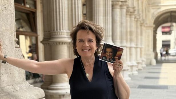 Frau im schwarzen Top und Leopardenhose steht in einer säulengesäumten Passage und hält eine CD hoch.