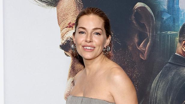 Sienna Miller lächelt vor einem Filmplakat in die Kamera.