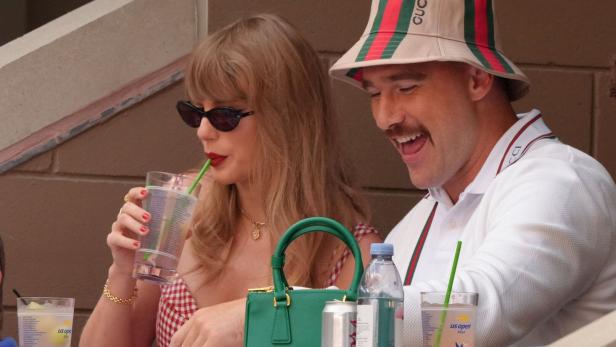 Taylor Swift und Travis Kelce sitzen nebeneinander bei den US Open, wobei Swift ein Getränk trinkt und Kelce lachend einen Gucci-Hut trägt.
