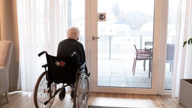 Ältere Person im Rollstuhl blickt durch die Terrassentür auf den Balkon.
