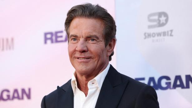 Dennis Quaid lächelt vor einem Hintergrund mit dem Schriftzug „Reagan“ in die Kamera.